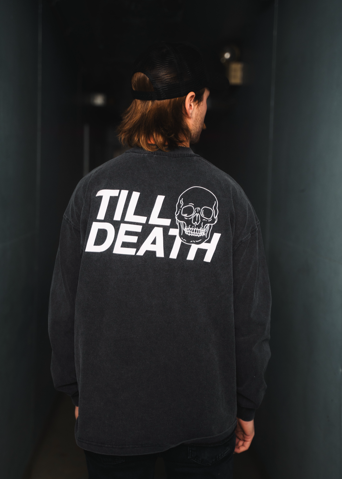 Person wearing a black long-sleeve shirt with 'Till Death' graphic on the back against a dark background